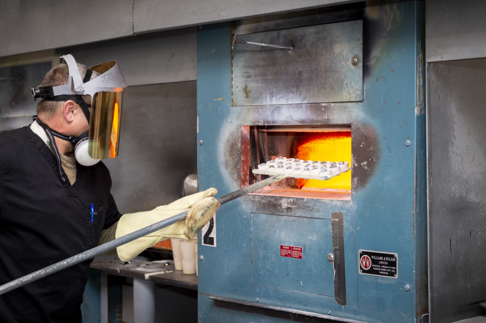 A worker in full protective equipment sliding a tray of crucibles into a glowing muffle furnace in the Eastern Analytical fire-assay lab