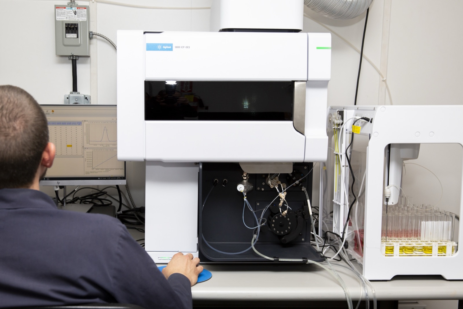 An analyst seated at a monitor running the ICP software beside the lab's ICP-OES instrument, with sample vials in an autosampler tray