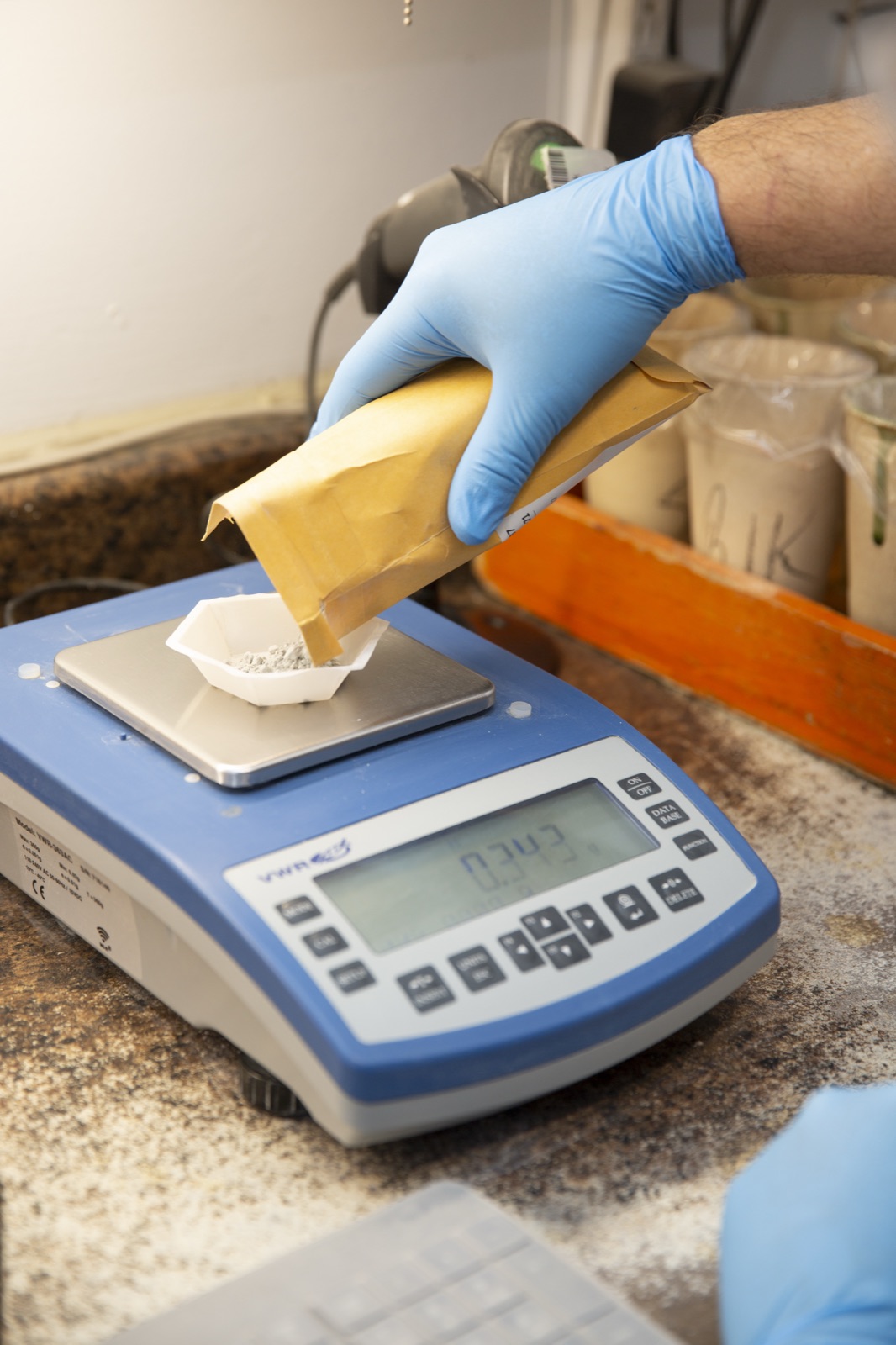 A gloved hand pouring pulverized sample from a kraft envelope into a weigh boat on a digital analytical balance